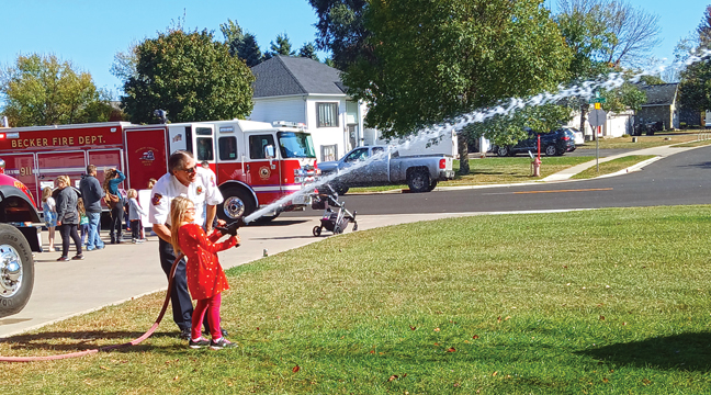 BEcker Firefighter Dave Daulhimer aided second grader Aspen Haas with the fire hose during last weekend’s BFD Open House. (Patriot Photo by Deven Haas).