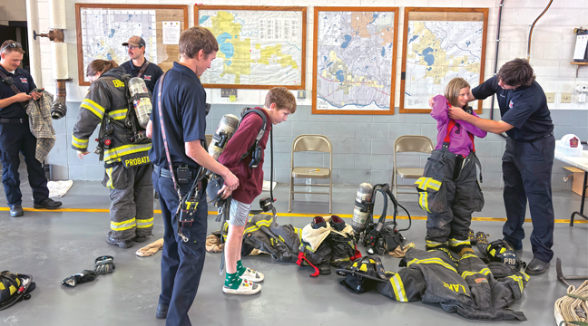 Firefighter Jesse Keller (L) watches wife Kendra put on gear while FF Jake Cihlar (back) offers advice.  FF Colin Maki (M) helps Parker Just and FF Jacob Dietrick (R) helps Annabelle Norlin.  (Patriot photo by Don Bellach)