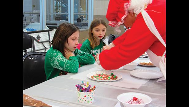 MRS. CLAUS DEMONSTRATES SOME BAKING TRICKS to Maggie and Ella Ackman. (Photo by Penny Leuthard)